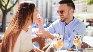 Ein junges Pärchen sitzt im Eiscafe. Der junge Mann bietet seiner Freundin sein Eis auf seinem Löffel zum probieren an.